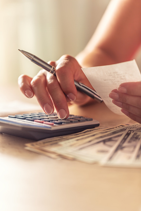 女性手中拿着一张旁边有美元纸币的钞票，手里拿着计算器的细节。图片下载